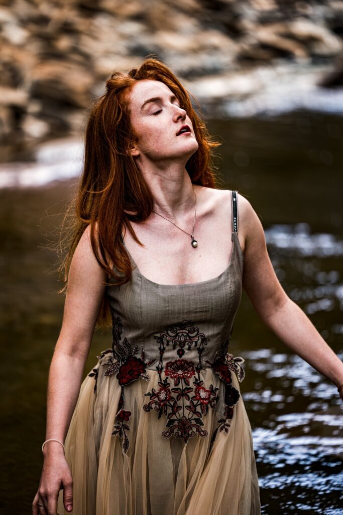 Model im handbestickten Kleid steht im Fluss, Blick zum Licht, gestickte Blumen in Rot und Schwarz an der Taille