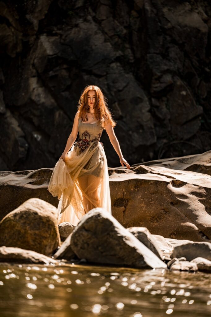 Model geht im Kleid über sonnenbeschienene Felsen am Wasser, der Tüll leuchtet im Gegenlicht