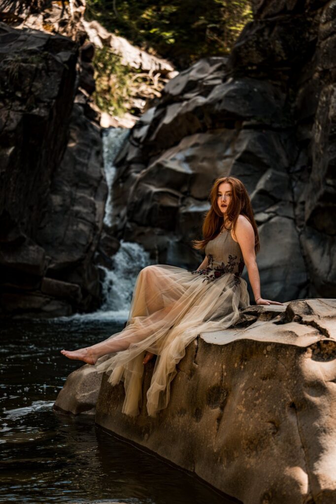 Model sitzt barfuss im Kleid auf einem Felsen am Wasserfall