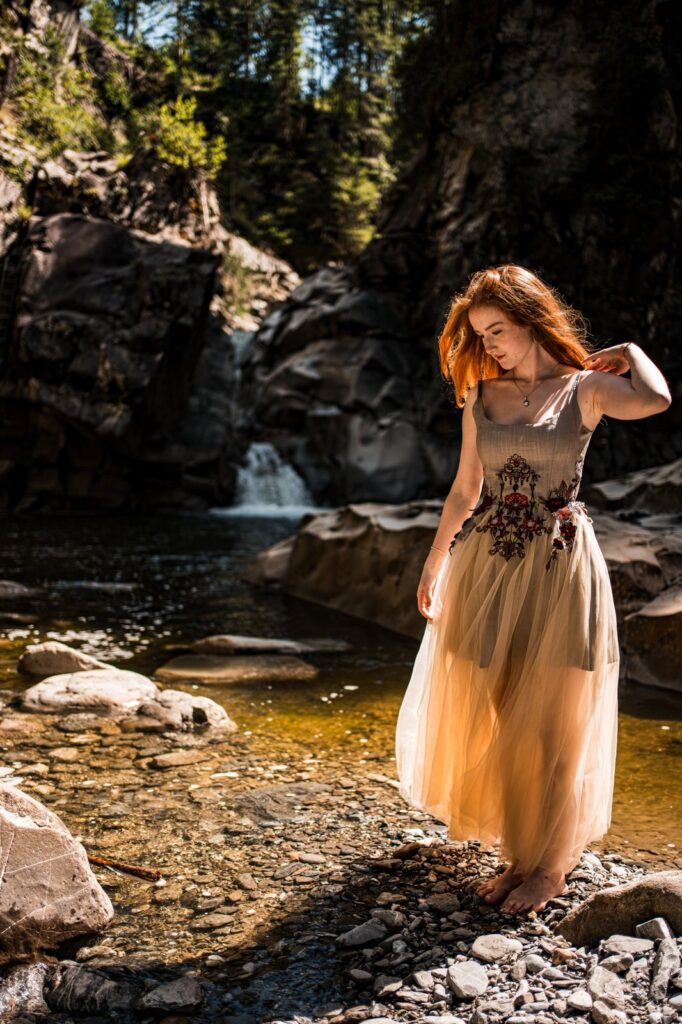 Model steht barfuss im Kleid am Ufer eines Bergbachs