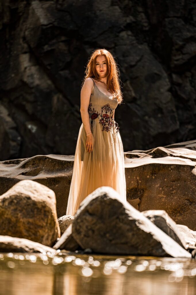 Model steht im Kleid zwischen Felsen, die gestickten Blumen an der Taille im Licht
