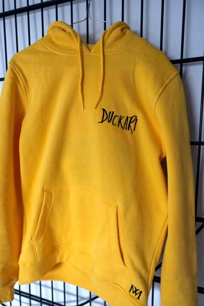 Gelber Duckari Hoodie an einem Gitter, Schriftzug auf der Brust und Monogramm am Saum