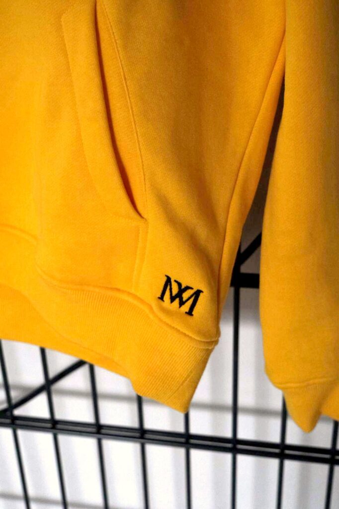 Gesticktes MM-Monogramm in Schwarz am Saum des gelben Hoodies