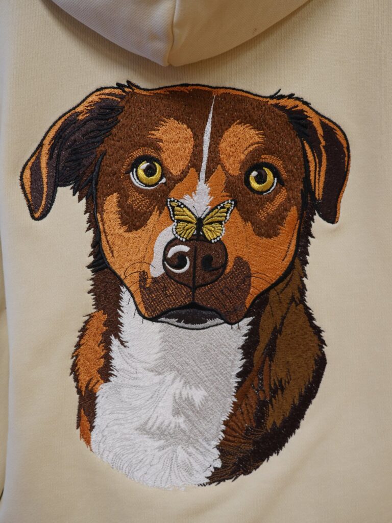 Gesticktes Hundeporträt auf dem Rücken eines cremefarbenen Hoodies, mit goldenem Schmetterling auf der Nase