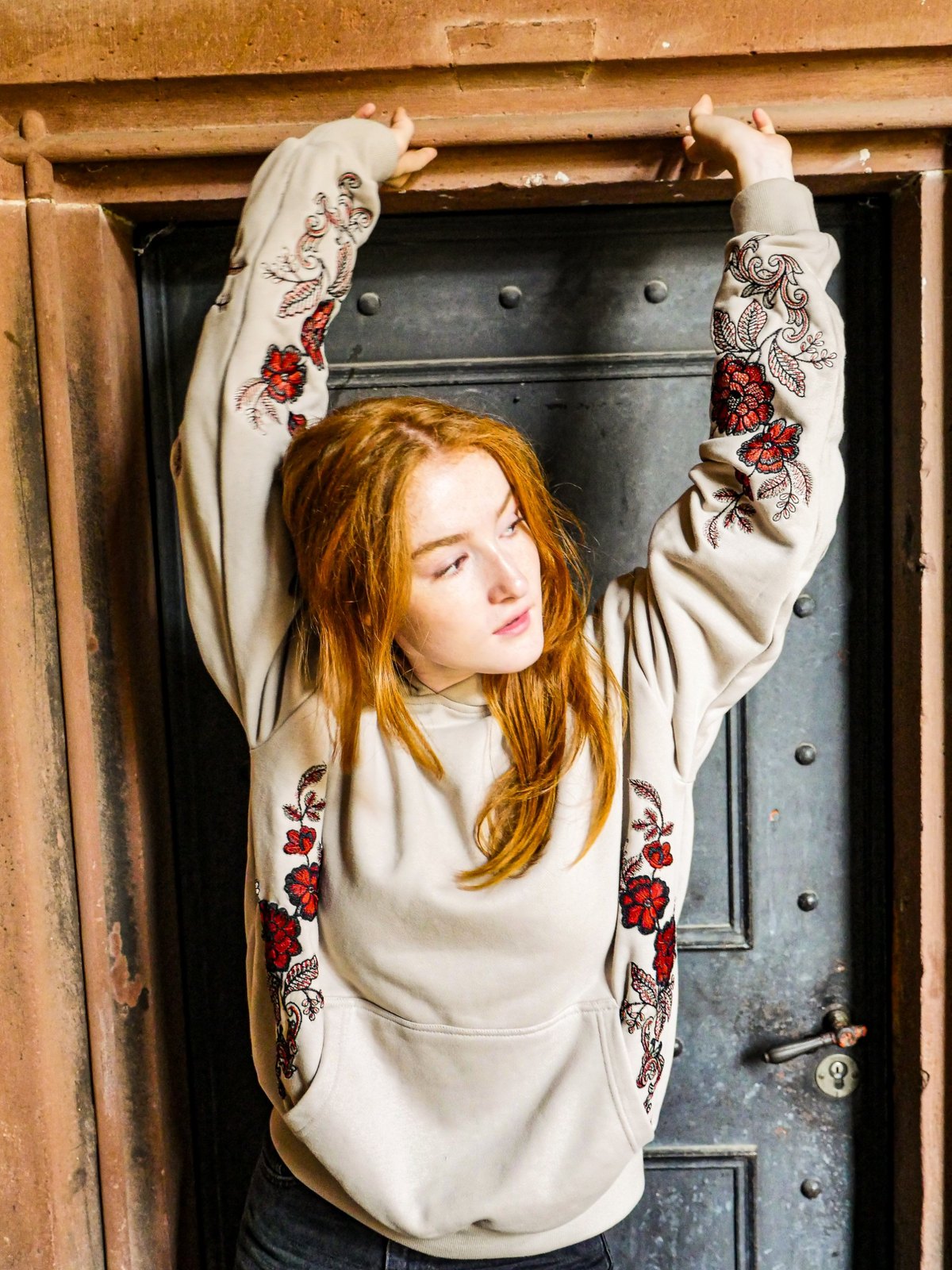 Model im sandfarbenen Oversize Hoodie mit gestickten Blumen an den Ärmeln, Arme über dem Kopf an einem alten Türrahmen