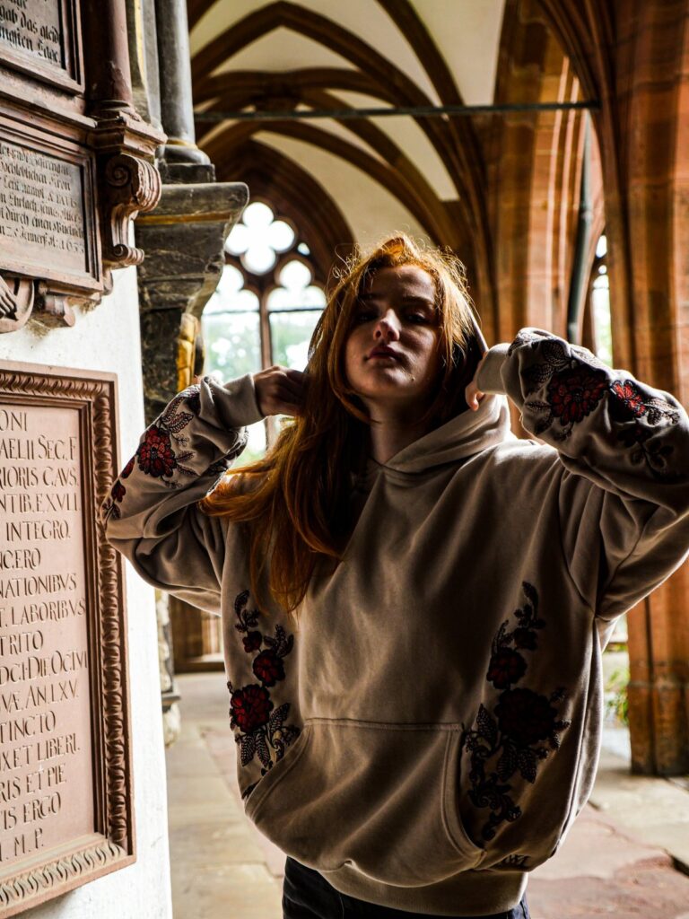 Model zieht die Kapuze des bestickten Oversize Hoodies hoch, im Kreuzgang mit gotischen Bögen