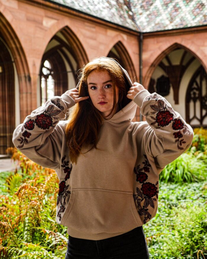 Model mit Kapuze im Innenhof vor gotischen Bögen, Blumenstickerei an Ärmeln und Seiten des Hoodies