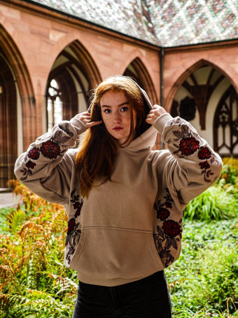 Model mit Kapuze im Innenhof vor gotischen Bögen, Blumenstickerei an Ärmeln und Seiten des Hoodies