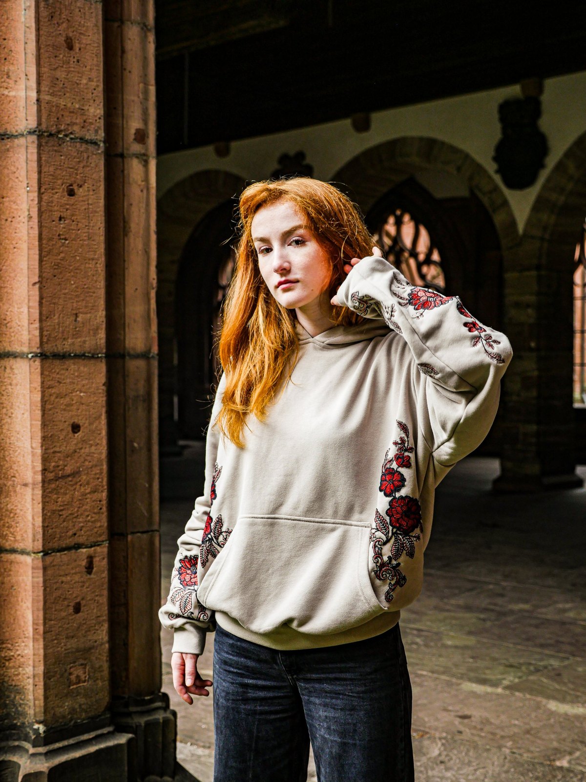 Model im sandfarbenen Oversize Hoodie mit gestickten Blumen, neben einer Sandsteinsäule